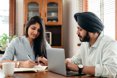 Man and woman working at a table. The woman is looking down towards the table whilst the man is looking towards her with a laptop in front of him, he's also wearing a headscarf.