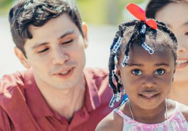 young girl looking into the camera with two adults behind her looking at her